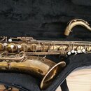 Zlatý saxofon v černém pouzdře. Povrch nástroje vykazuje výrazné opotřebení, škrábance a oděrky laku. Na instrumentu nejsou viditelné žádné značky an…