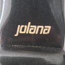 Detail černého lakovaného povrchu hudebního nástroje. Uprostřed je bílý nápis jolana. Pod nápisem jsou viditelné jemné rýhy a drobné nečistoty v laku.