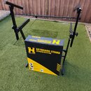 Černý kovový stojan na klávesy v rozloženém stavu. Uvnitř stojanu se nachází žlutý a černý kartonový box s nápisy HERCULES., KEYBOARD STAND KS400B/KS…
