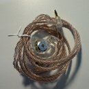 Sluchátka in-ear s průhledným pláštíkem a pleteným kabelem v měděné barvě. Na vnitřním kovovém prvku sluchátka je viditelné logo ve tvaru trojúhelník…