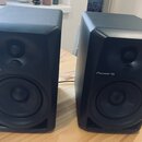 Dva černé studiové reproduktory s hranatým tvarem. Na přední straně každého reproduktoru je bílý nápis Pioneer DJ, nadstavba s výškým reproduktorem a…