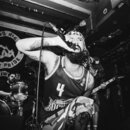 Černobílá fotografie zachycuje zpěváka s mikrofonem v ruce a bubeníka za ním. V pozadí je vidět vlajka s nápisem GOOD NIG WHITE PRIDE a adrese WWW.GI…