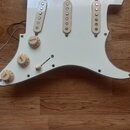 Bílá plechová vrstva (pickguard) pro elektrickou kytaru s třemi bílo-krémovými snímači typu single coil. Na knoflících jsou viditelné nápisy VOLUME a…