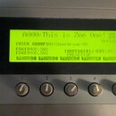 Detailní záběr na ovládací panel elektronického hudebního zařízení s šedým povrchem. Uprostřed je umístěn žlutý displej s textem: „A000:This is Zee O…