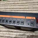 Černý napájecí adaptér s oranžovými popisky. V levé části je sekce DC 9V OUT se sedmi výstupními konektory, nad nimi jsou nápisy 100mA (5x) a 250mA (…