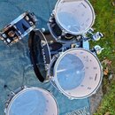 Sada černých bubnů značky YAMAHA s kovovými obručmi. Na fotce jsou vidět čtyři bubny: snare, tom a dva basové/floor tomy. Na bílých blanách je čiteln…