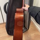 Zadní strana akustické kytary s hnědým lakovaným povrchem z dřeva. Nástroj je umístěn před otevřený černý softcase. Na fotce nejsou žádné viditelné z…