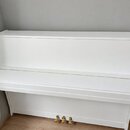 Bílé piano s uzavřenou klaviaturní víkem. Nástroj má hladký bílý povrch a tři zlaté pedály v dolní části. Na viditelných místech nejsou žádné značky…