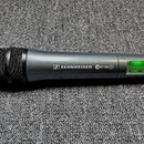 Šedý bezdrátový mikrofon s černou mřížkou a fialovým prstencem u spodní části. Na těle je bílý nápis SENNHEISER s logem a označení ew 100 G2. V zadní…