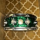 Zelený buben s kovovými obručmi a mechanismy. Uprostřed korpusu je kulatý štítek s nápisy „DW“ a „Drum Workshop“. Povrch nástroje je v dobrém stavu b…