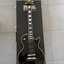 Černá elektrická kytara s bílým lemováním, položená na černém kufru s nápisem PAUL REED SMITH GUITARS. Na hlavě kytary je zlaté logo s nápisem Paul R…