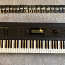 Černý klavír s nápisy KURZWEIL a K2000 V2. Nástroj má ovládací panel s displejem, knoflíkem a řadou tlačítek. Nad klavírem je v řadě rozloženo 24 dis…