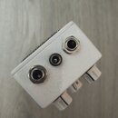Bílá kytarový efekt pedál s kovovým šasi. Na zadní straně jsou dva 6,3mm jack konektory a jeden malý napájecí vstup. Na horní hraně je viditelná část…