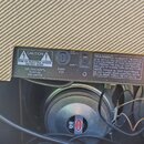 Zadní strana zesilovače s tkanou látkovou krytkou. Na kovovém panelu jsou varovné nápisy CAUTION RISK OF ELECTRIC SHOCK DO NOT OPEN a WARNING: TO RED…
