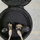 Černé sluchátka in-ear v kulatém černém pouzdře. Na pravém sluchátku je viditelný nápis se846 a logo Shure. Sluchátka mají průhledné kabelové zakonče…