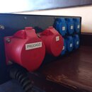 Černý rozvodnicový box s několika elektrickými zásuvkami. V popředí jsou dvě červené zásuvky, z nichž jedna má bílou nálepku s nápisem PRŮCHOZÍ a dru…