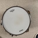 Fotografie zachycení bubnu z ptačí perspektivy. Na bílém bláně jsou vidět nápisy REMO AMBASSADOR COATED MADE IN U.S.A. a TAMA. Kovový obruč je stříbr…