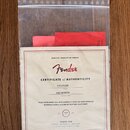 Průhledný plastový sáček s dokumenty, v němž je vidět bílý certifikát s červeným rámem. Na certifikátu je logo Fender a nápis CERTIFICATE of AUTHENTI…