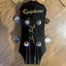 Černá hlava kytary s nápisem Epiphone v horní části a Les Paul MODEL v dolní části, oboje provedená zlatým písmem. Jsou vidět čtyři kovové ladičky a…