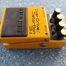 Žlutý kytarový efekt s černým nášlapným spínáčem. Na šasi je nápis BOSS a OverDrive/ Distortion OS-2. Přístroj má čtyři žluté ovládací knoflíky a zna…