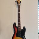 Elektrická basová kytara s tělem v provedení sunburst a černým pickguardem. Na hlavě je nápis JAZZ BASS. Nástroj má hnědý hmatník s bílými polohovými…