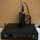 Černý bezdrátový přijímač s dvěma anténami a připojeným vysílačem. Na přední straně přijímače jsou nápisy AUDIO OUTPUT, BALANCED, UNBALANCED, SQUELCH…