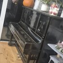Černý pianino s ornamentálními vzory na přední části. Povrch nástroje vykazuje viditelné opotřebení, škrábance a oděrky. Na instrumentu nejsou čiteln…
