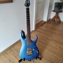 Modrá elektrická kytara s kovovými zlatými komponenty, umístěná na černém stojanu. Na hlavě je viditelné logo Ibanez. Kytara má dva humbuckery, tremo…