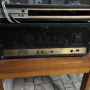 Černý kytarový zesilovač v koženém potahu s kovovou mřížkou. Na zlatavém ovládacím panelu je nápis ATTACK POWER HEAVYMASTER a několik ovladačích knof…