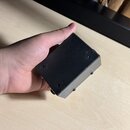 Černý kovový box pravděpodobně sloužící jako efekt pedál nebo napájecí adaptér. Povrch je matný s viditelnými známkami opotřebení a drobnými odřapkán…