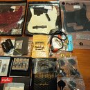 Sada komponentů pro kytaru rozložená na dřevěném stole. V horní části jsou tři pickguardy v plastových obalech s nápisem FLEOR, jeden v bordové barvě…