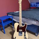 Světle béžová elektrická kytara s tmavě hnědým pickguardem, umístěná na černém stojanu. Na hlavě nástroje je viditelné logo Fender. Kytara má šest st…
