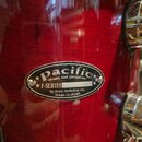 Detail červeného bubnového korábu s lakovaným povrchem. Na nástroji je oválný kovový štítek s nápisy „pacific drums and percussion“, „F-SERIES“ a „by…