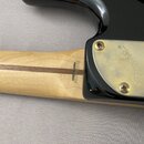 Detail krku a spoje s tělem černé kytary. Na zlatém kovovém krytu je vyryto logo Fender®. Na dřevěném krku je černý nápis MADE IN JAPAN, pod kterým j…