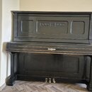 Černý piano v přímém záběru zepředu. Nástroj má zdobný ornament uprostřed klaviaturního víka a tři mosazné pedály. Povrch vykazuje známky opotřebení…
