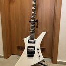 Bílá elektrická kytara s ostře hranatým tvarem těla a špičatou hlavou. Na hlavě je viditelné logo Jackson. Kytara má dva černé humbuckery, dvě ovláda…