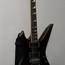 Černá elektrická kytara s ostře hranatým tělem, umístěná na nástěnním držáku. Na hlavě je viditelné logo Ibanez. Kytara má dva humbuckery, tremolo mo…