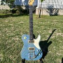 Elektrická kytara s modrým třpytivým povrchem těla a bílým pickguardem. Na hlavě hředu je viditelné logo Fender. Kytara má dva humbuckery, jeden ovlá…