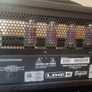 Zadní strana kytarového zesilovače s černou mřížkou, za kterou jsou vidět čtyři elektronky a kondenzátory. Na spodním černém panelu je bílý štítek s…