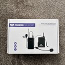 Bílé papírové balení bezdrátového mikrofonu. Na krabici je nápis MGZOS MIC-UHF-SET a WIRELESS MICROPHONE NOISE REDUCTION AND ANTI-INTERFERENCE. Upros…