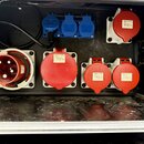 Vnitřek černého rozvodového kufru s několika elektrickými zásuvkami. V horní části jsou tři modré zásuvky a jedna červená zásuvka s nápisem INDU-ELEC…