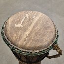 Djembe s tělem z tmavého dřeva a napnutou kůží v horní části. Okraj bubnu je opleten zeleným šňůrkem a spodní část je zajištěna černými provazky. Na…