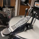 Krytý basový pedál s kovovou konstrukcí a stříbrným šlapadlem. Na horní části je nápis The Drummer's Choice,® a čtyři oválné otvory. Pedál je umístěn…