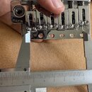 Detailní záběr na chromovaný kovový kobylcový systém kytary, který je změřován digitálním posuvným měřidlem. Na levé straně je viditelné logo „LOK“.…