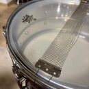 Detailní pohled na snare drum s průhlednou blánou značky REMO. Na bláně je položen kovový sítěný mechanismus (strained) s nápisem Ludwig. Kovové obru…