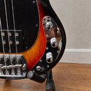 Detail elektrické basové kytary s černým pickguardem a tělem v červeno-žlutém sunburst provedení. Na chromovaném ovládacím panelu jsou tři kovové oto…