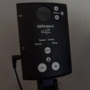 Černý ovládací modul elektronických bicích s nápisy Roland a V-Drums. Na panelu jsou tlačítka Volume, Select, plus (+) a minus (-), oraz nápisy Tempo…