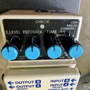 Kytarový efekt pedál s krémovým šasi a černým ovládacím panelem. Na panelu jsou čtyři černé potenciometry s modrými knoflíky a nápisy E.LEVEL, FEEDBA…