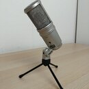 Stříbrný kondenzátorový mikrofon s kovovou mřížkou, umístěný na malém černém stativu. Na těle mikrofonu je nápis E205U a v dolní části značka CE. Na…