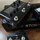 Černý efektový pedál s nápisem LINE 6 HX STOMP, vybavený displejem, několika ovládacími knoflíky a třemi stoupakovými spínači. Nástroj leží na černé…